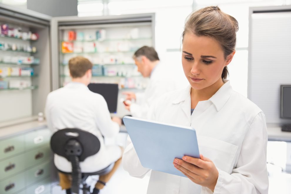 just-in-time-en-sanidad Mujer en bata blanca en el entorno de un gabinete farmacéutico o médico con tableta electrónica para ilustrar el concepto just in time en sanidad